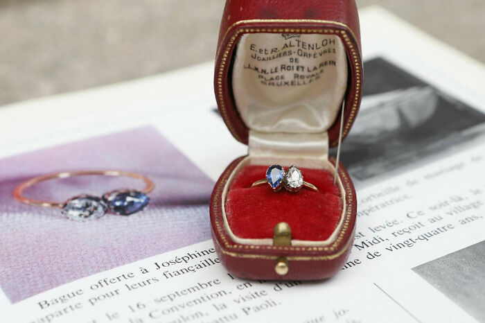 A vintage ring with gem settings in an open red velvet box, placed on a historical book page of artifacts.