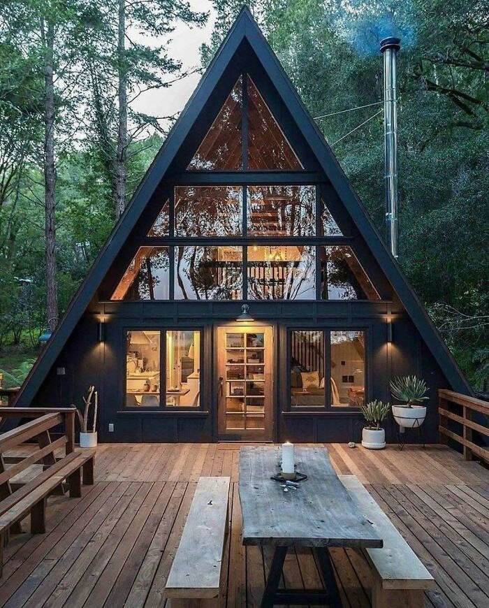Modern A-frame tiny home with large windows and wooden deck surrounded by trees, inspiring life off the grid.