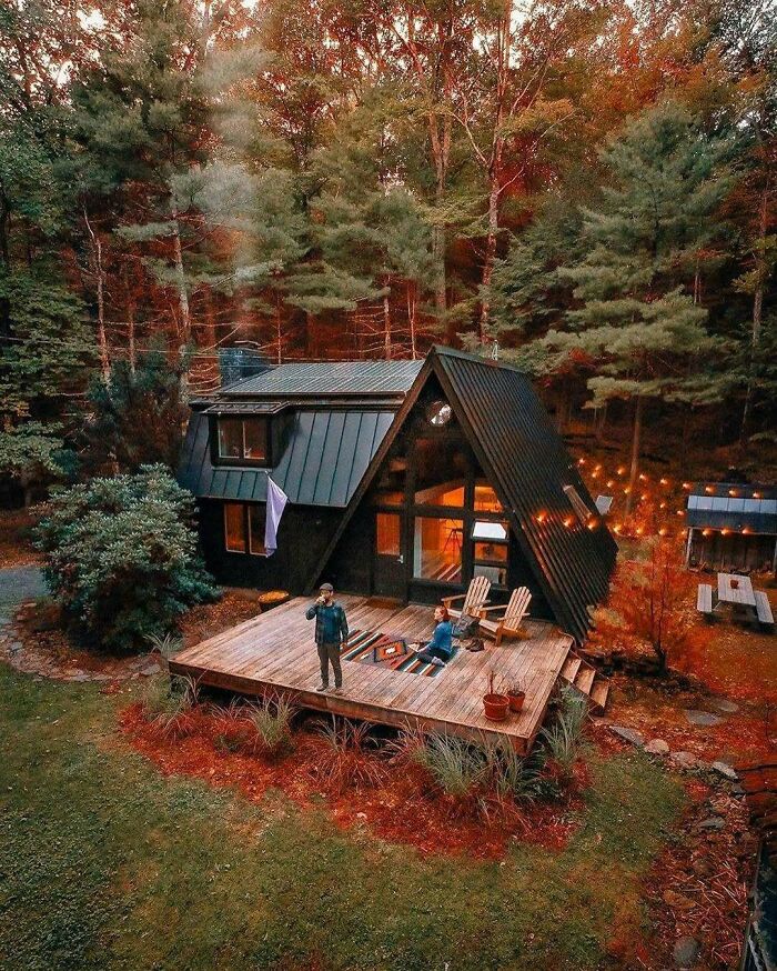 Modern tiny home with A-frame design surrounded by autumn forest, featuring a wooden deck and outdoor seating area off the grid.
