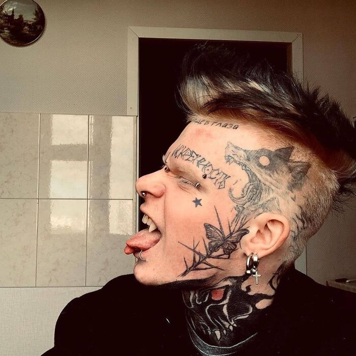 Person with unusual tattoos covering face and neck, showcasing a wolf and other designs in a bathroom setting.