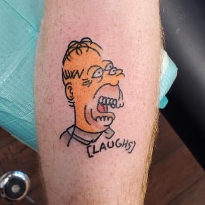 Tattoo of a distorted Homer Simpson on a leg, with "LAUGHS" text below.