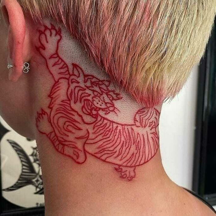 A hilariously bad tattoo of an outline of a tiger in red ink on the side of a person's neck, under short blonde hair.