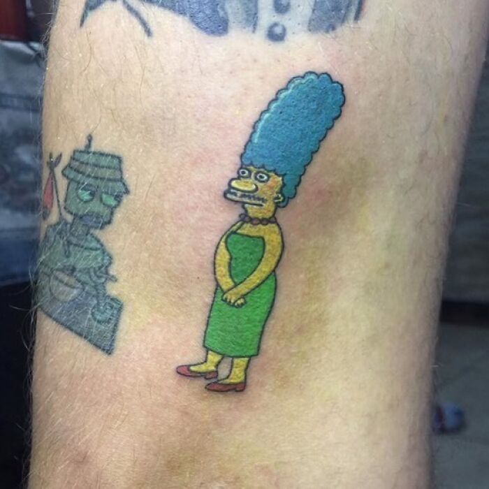 A Simpsons tattoo of Marge with blue hair and a green dress on a person's skin.