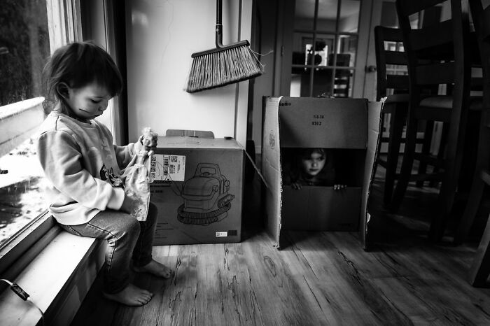 Two children playing in boxes indoors, one sitting by a window and another peeking out, capturing rural life in Maine.