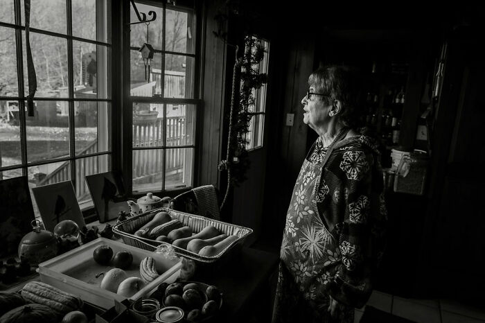 Woman gazing out a window in rural Maine, surrounded by fresh produce, capturing intimate rural life.