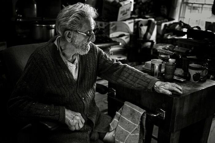 Elderly man in a cozy room, symbolizing intimate rural life in Maine, seated next to a table with personal items.