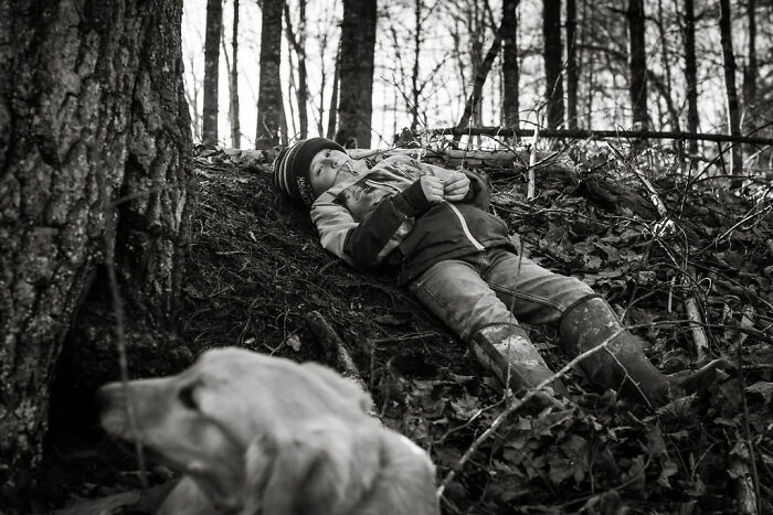 Child in rural Maine resting against a tree, lying on the ground in a winter coat, with a dog in the foreground.