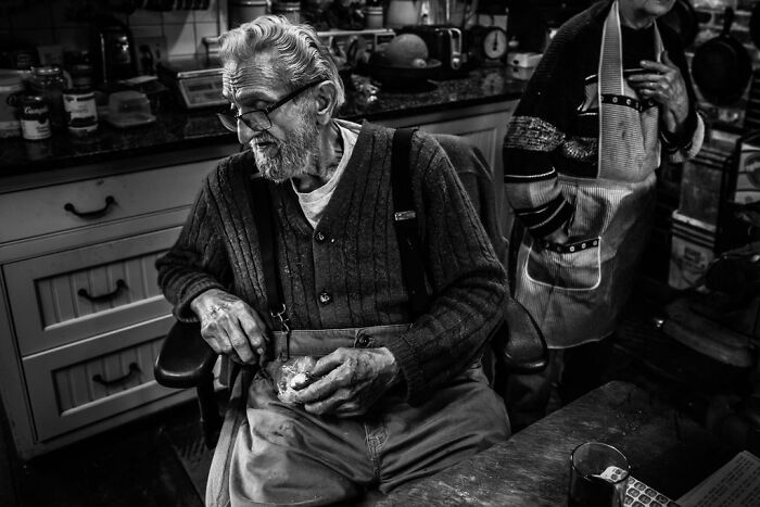 Elderly man in a rural Maine kitchen peeling an apple, wearing glasses and a cardigan, capturing intimate rural life.