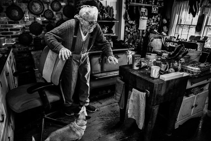 Elderly man in rural Maine kitchen, interacting with a small dog, surrounded by rustic decor and a wood stove.