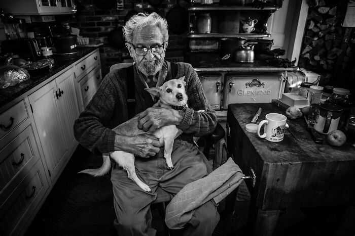 Elderly man in rural Maine kitchen holding a small dog, showcasing intimate rural life.