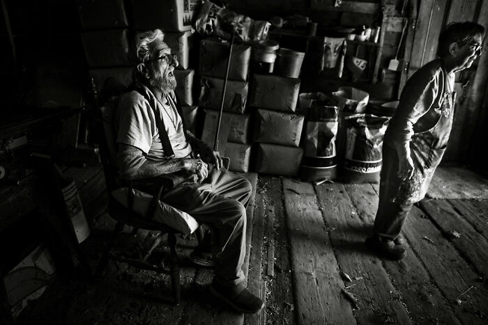 Elderly man sitting and woman standing in a rustic room, capturing rural life in Maine through intimate photography.