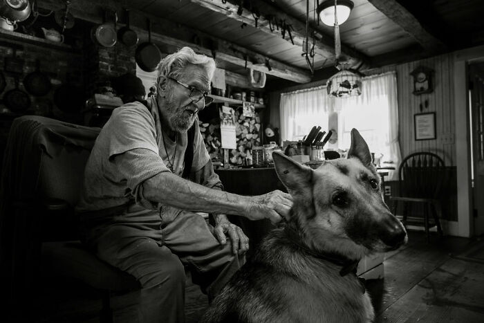 Elderly man in rustic Maine home with German Shepherd, capturing rural life's intimacy.
