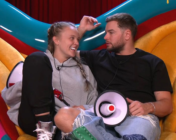 Two people sharing a close moment on a colorful couch in a Big Brother setting with megaphones nearby.