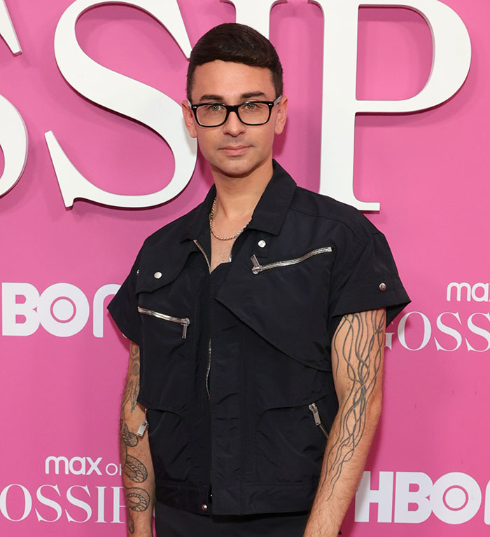 Man in black outfit with tattoos, wearing glasses, stands against a pink backdrop at a red carpet event.