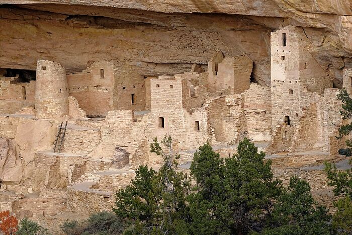 Ancient stone structures in an eerie underground city nestled within a rocky cliff, surrounded by trees.