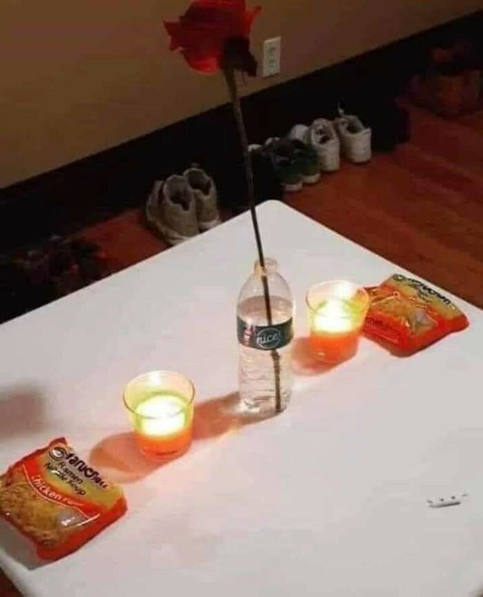 Unappetizing romantic dinner setup with instant noodles, bottle vase, and candlelight on a plain table.