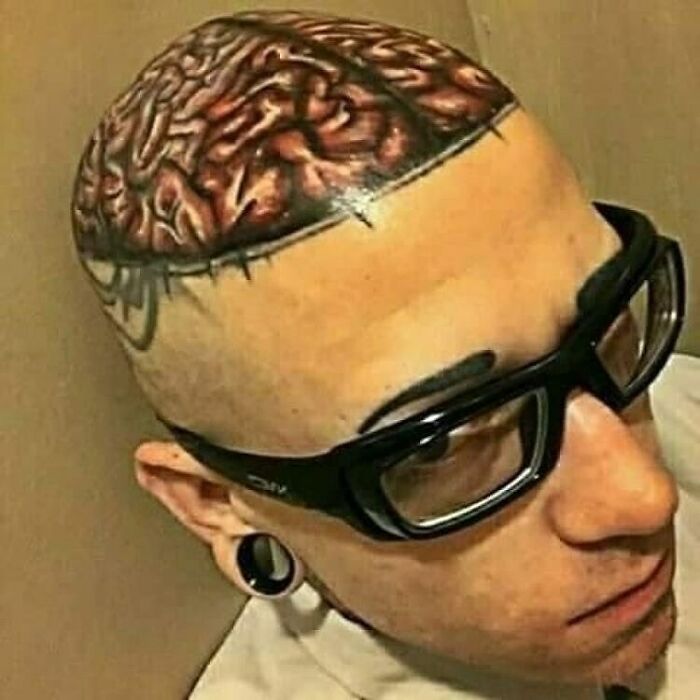 Person with a tattoo resembling a brain on their shaved head, wearing glasses and ear gauges.