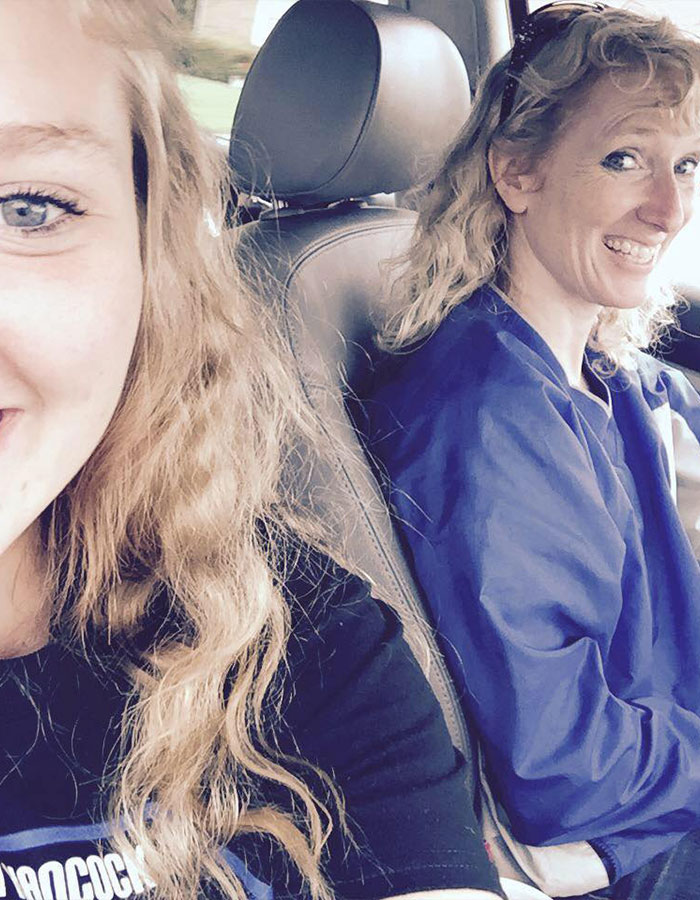 Two smiling women sitting in a car, one capturing a selfie, both with curly blonde hair.