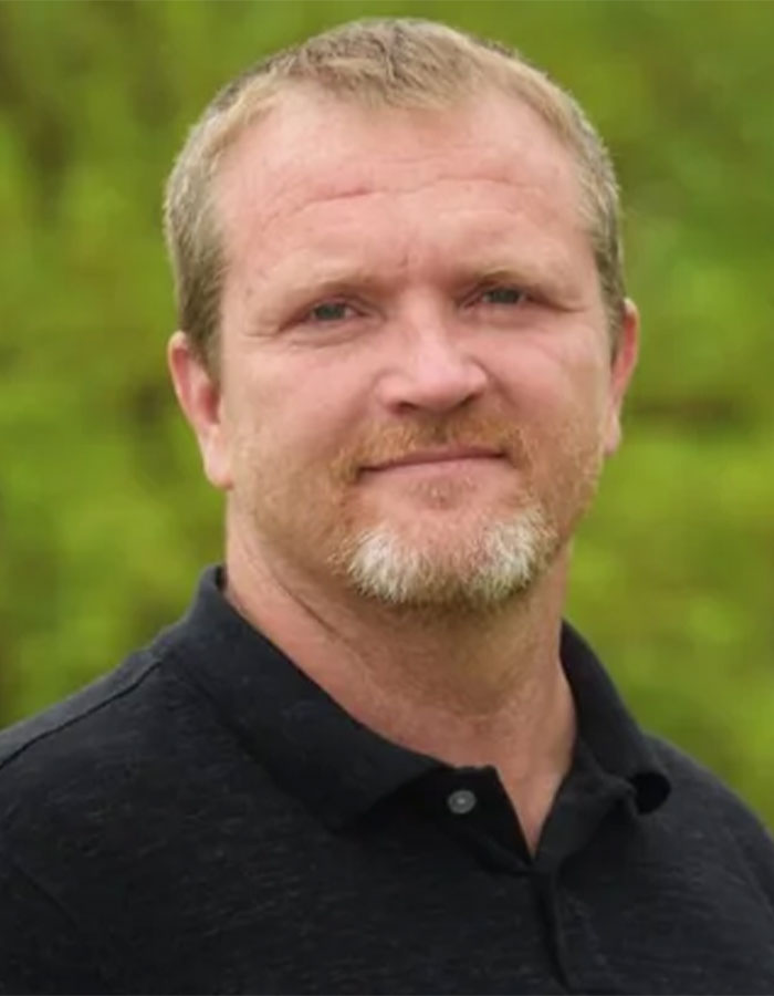 A man in a black shirt photographed outdoors with a blurry green background.