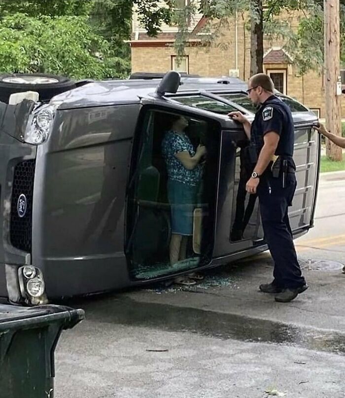 A police officer talks to a person inside a car tipped on its side, capturing a random cursed image.