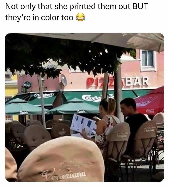 Couple at a cafe laughing over printed color memes about men, seated under an umbrella with "Pizza Bar" sign in the background.