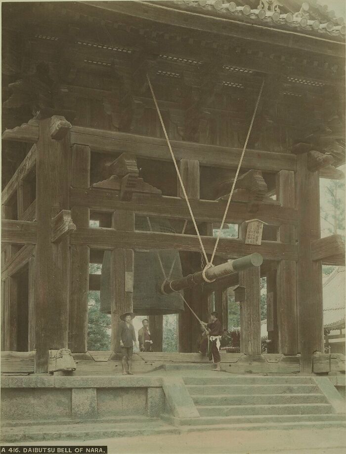 Large wooden bell structure with people standing nearby in a historical scene showing Japan in the 1800s.