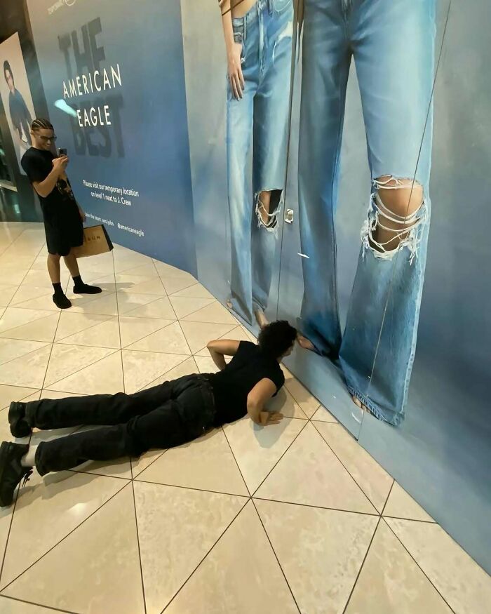 Person lying on a mall floor looking under a wall ad, with another person nearby on their phone.