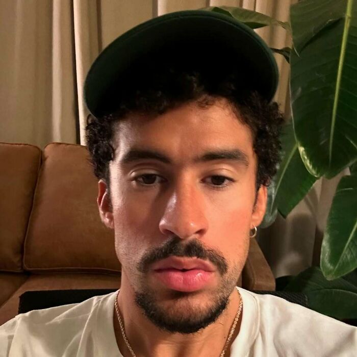 Celebrity in a casual setting, wearing a cap and white shirt, sitting on a couch with plants in the background.