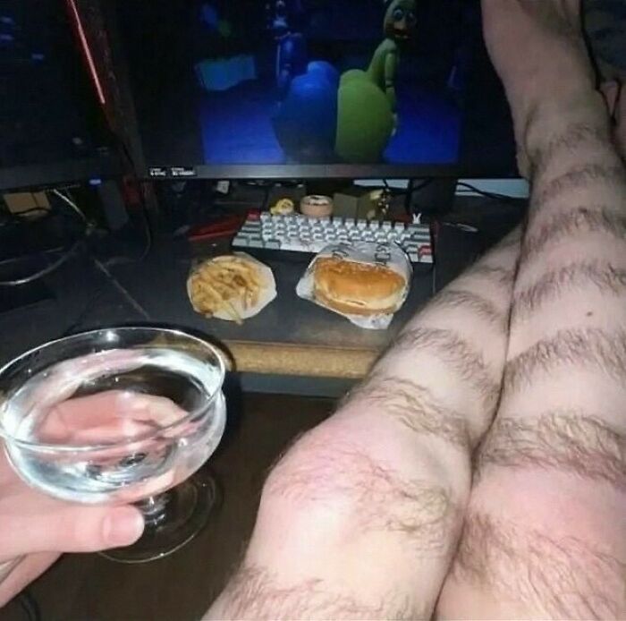 Person with hairy legs relaxes in front of a computer, holding a glass, with food items nearby from a popular IG page post.