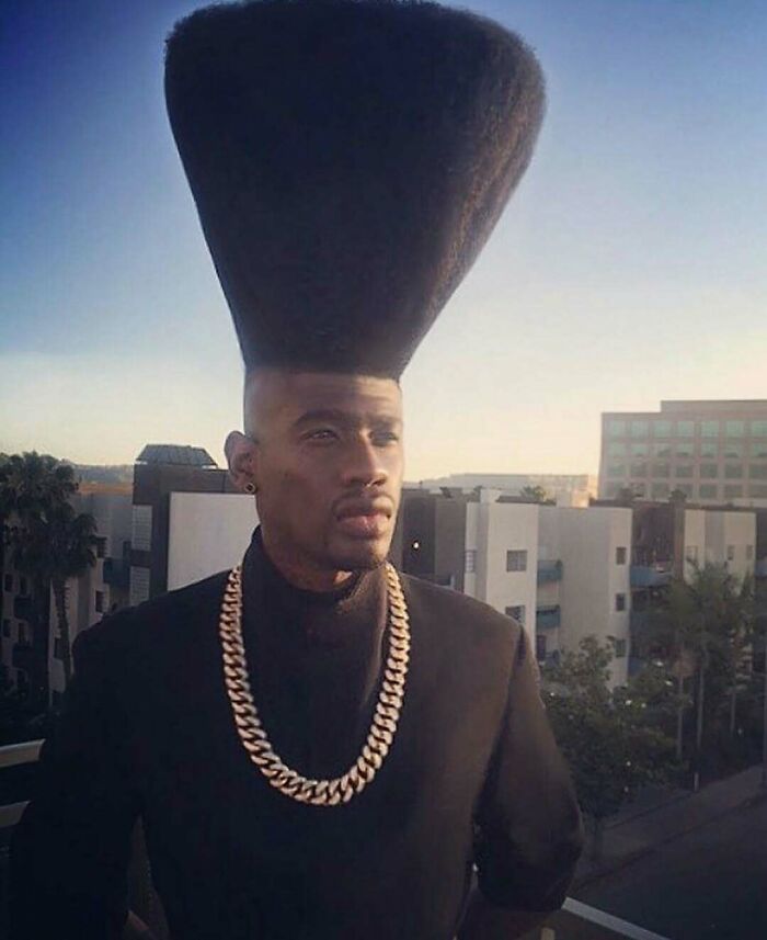 Man with towering hairstyle and gold chain standing on rooftop at sunset.