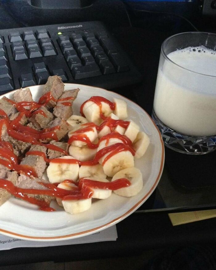 Plate of bananas and meat with ketchup, next to a glass of milk on a desk.