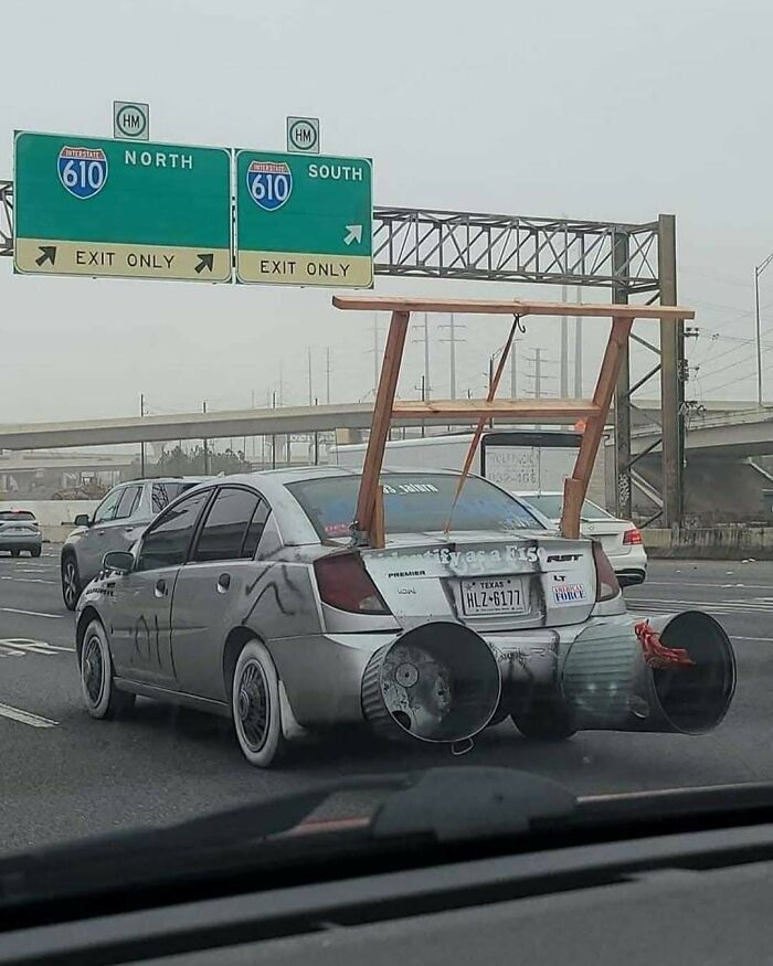 A car with humorous modifications and oversized exhausts driving on a highway under road signs. Car memes humor.