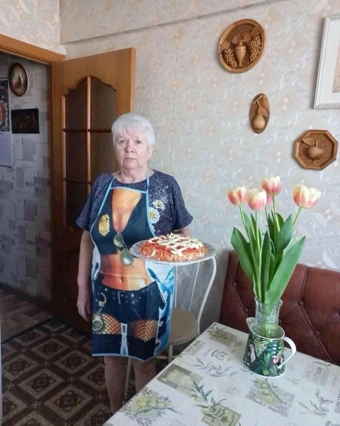 Elderly woman wearing a humorous apron holding a pizza, standing beside a table with tulips, showcasing unique IG content.