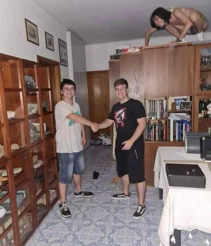 Two people shaking hands in a room while a third person crouches on a cabinet above, creating a random cursed image.