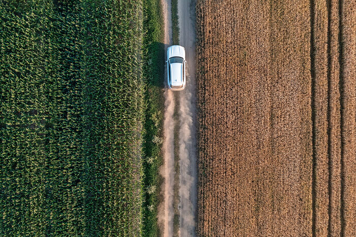 Aerial image of a car driving on a dirt road between green and brown fields, showcasing the world from above.