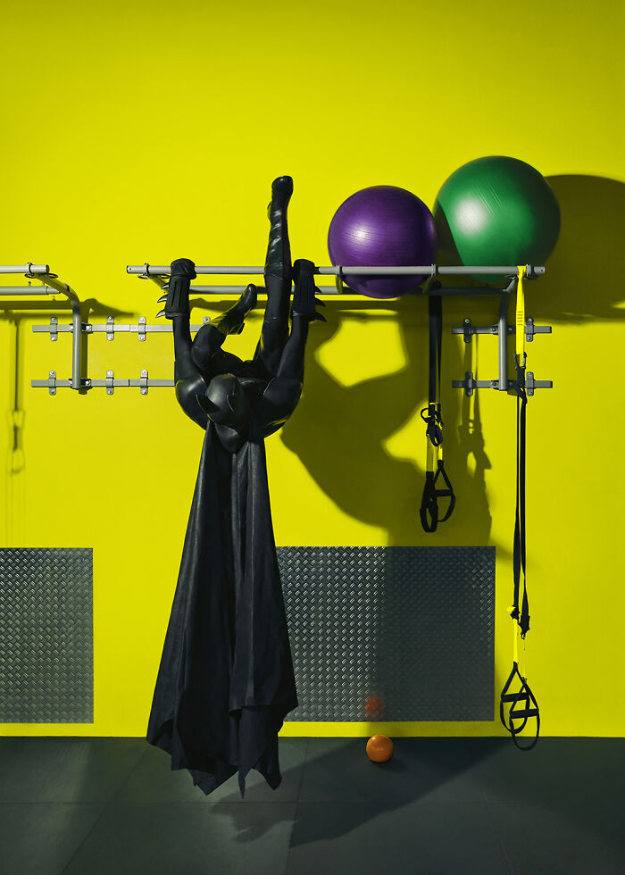 Batman exercising indoors, hanging upside down with gym equipment against a bright yellow wall.
