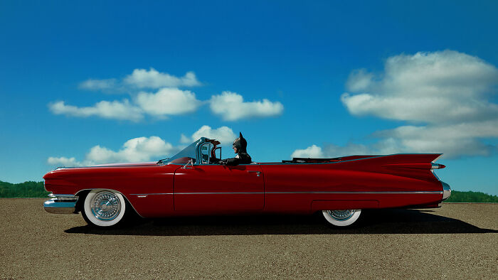 Batman in a red classic convertible driving under a blue sky, capturing a glimpse of normal life.