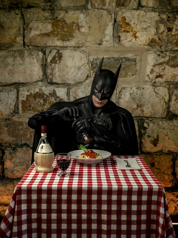 Batman dining alone, pouring wine over a pasta dish at a restaurant with a red-checkered tablecloth.