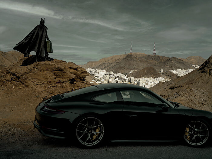 Batman stands on a hill with a suitcase, overlooking a cityscape and a parked black car in the foreground.
