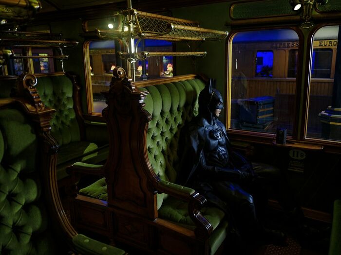 Batman sitting on a vintage train seat, surrounded by green upholstery and looking out the window.