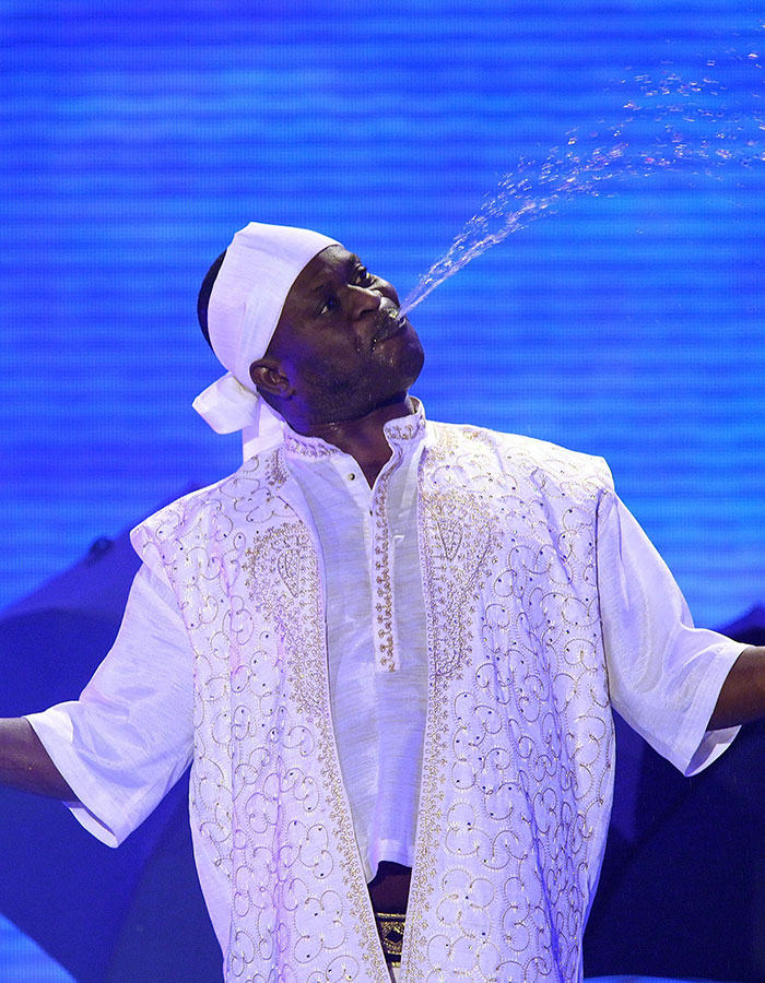 Man showcasing real-life superpowers by spitting water with precision, wearing traditional embroidered attire on stage.