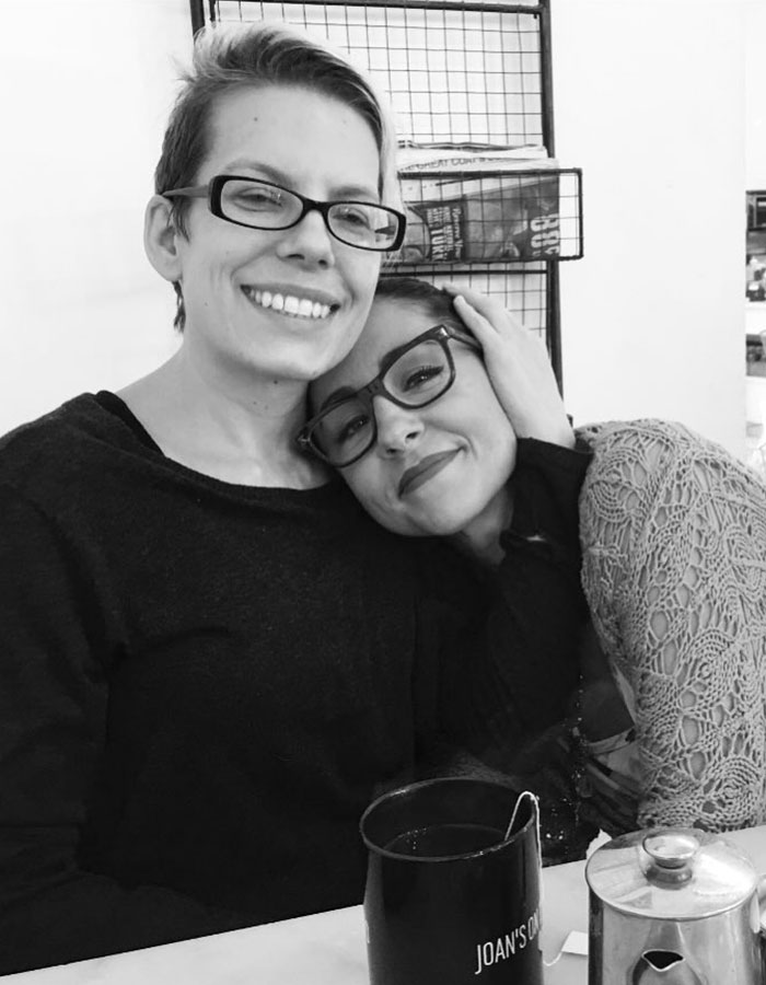 Two women smiling and embracing in a cafe, with a mug labeled "Joan's" on the table in front of them.