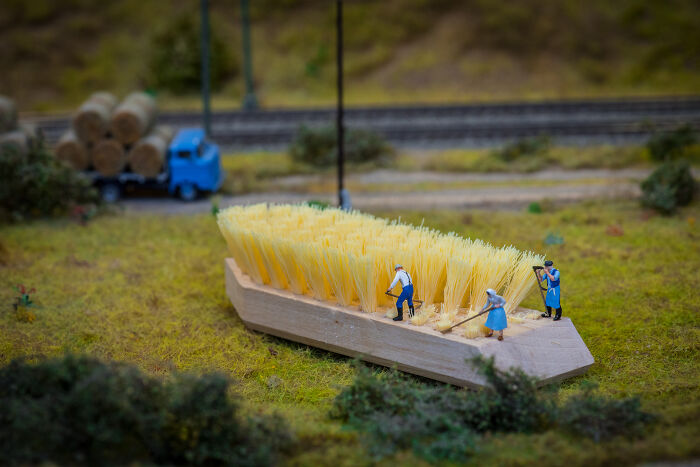 Miniature scene with tiny figures working on a yellow brush, resembling a field, with a model truck and logs in the background.