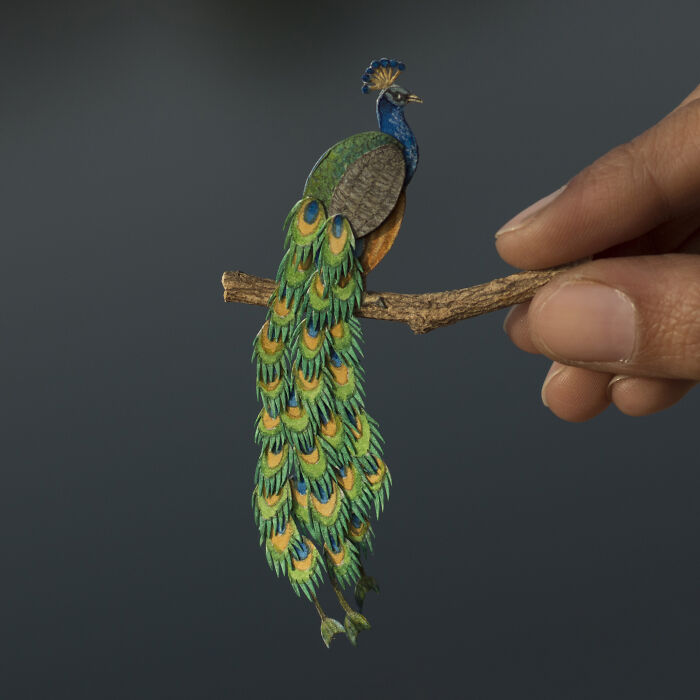 Miniature peacock sculpture on a small branch, intricately detailed, being held by fingertips.