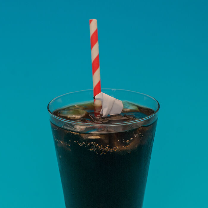 Miniature paper boat in a glass of soda, floating next to a striped straw, creating a secret world scene.