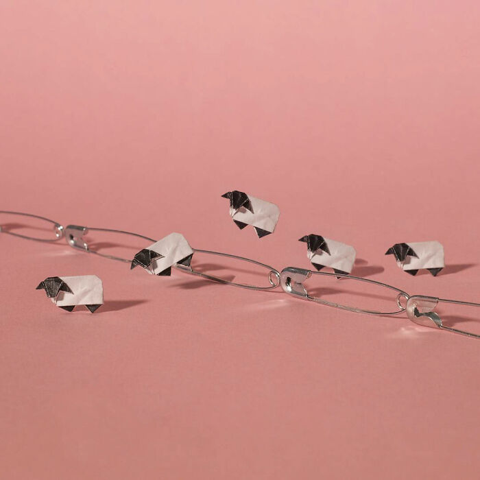 Miniature sheep crafted from paper perched on intertwined paperclips against a soft pink background.