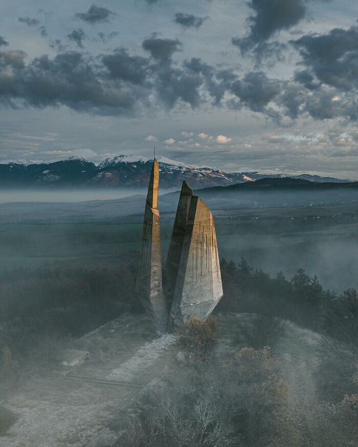 Drone photograph of a dramatic stone monument surrounded by mist and mountains in the background.
