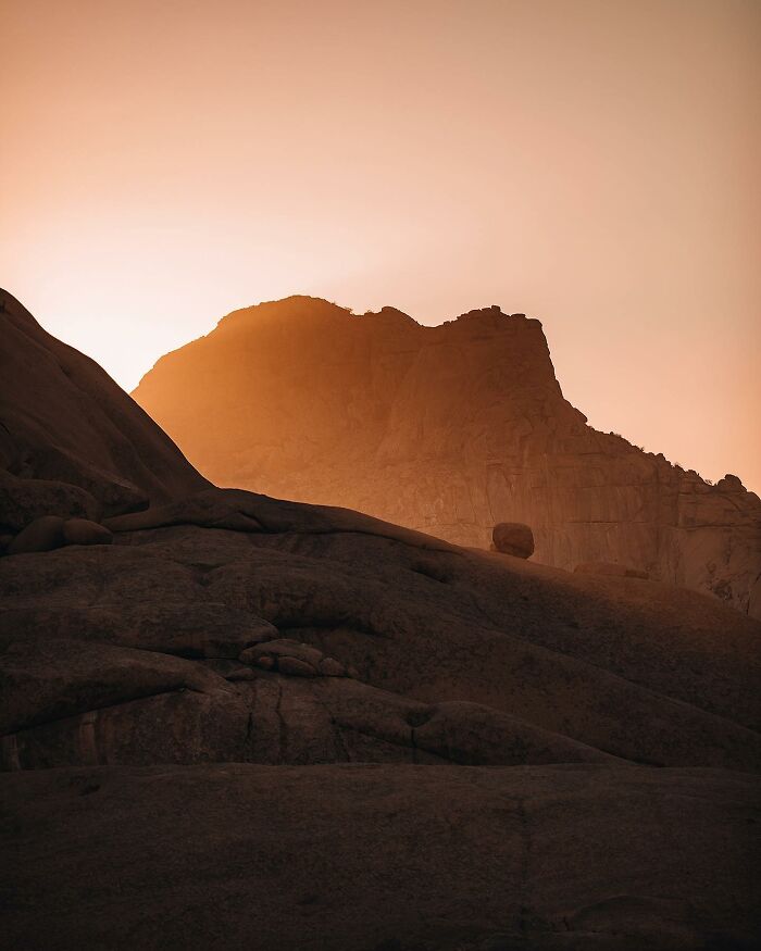 Sunset over rocky terrain, captured by drone, casting a warm glow over a mountain landscape.