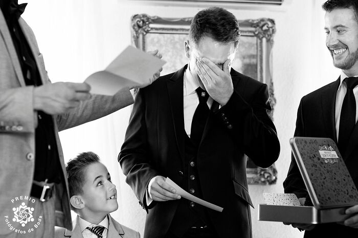 Groom wipes tears while holding vows, surrounded by smiling groomsmen and a young boy, at Premios FDB wedding photo awards.