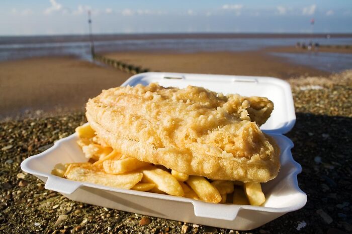 Fish and chips served in a takeaway box on a beach, showcasing a top dish from different countries.